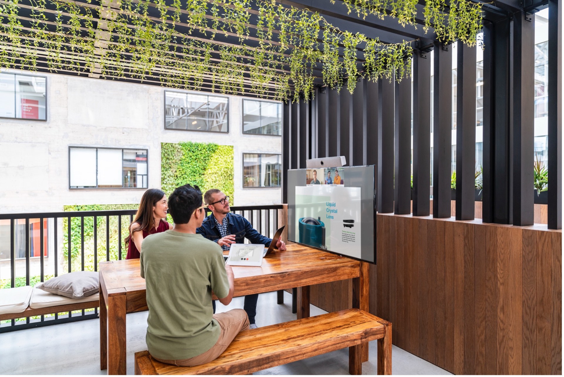 Three colleagues at a wooden table on a plant-covered terrace, joining a video call on a wall-mounted screen